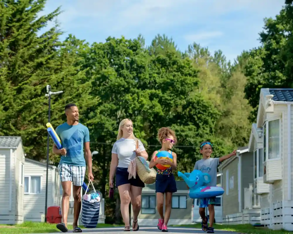 A family of four walks down a path lined with caravan homes. Two children hold inflatable pool toys, while an adult carries beach bags. The group smiles as they enjoy a sunny day, surrounded by lush green trees.