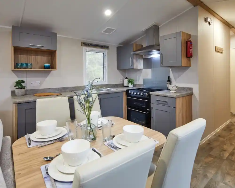 A modern kitchen interior featuring gray cabinets, a stainless steel oven, and a countertop with a small vase of flowers. A dining table set for four is in the foreground, with white bowls and glasses neatly arranged.