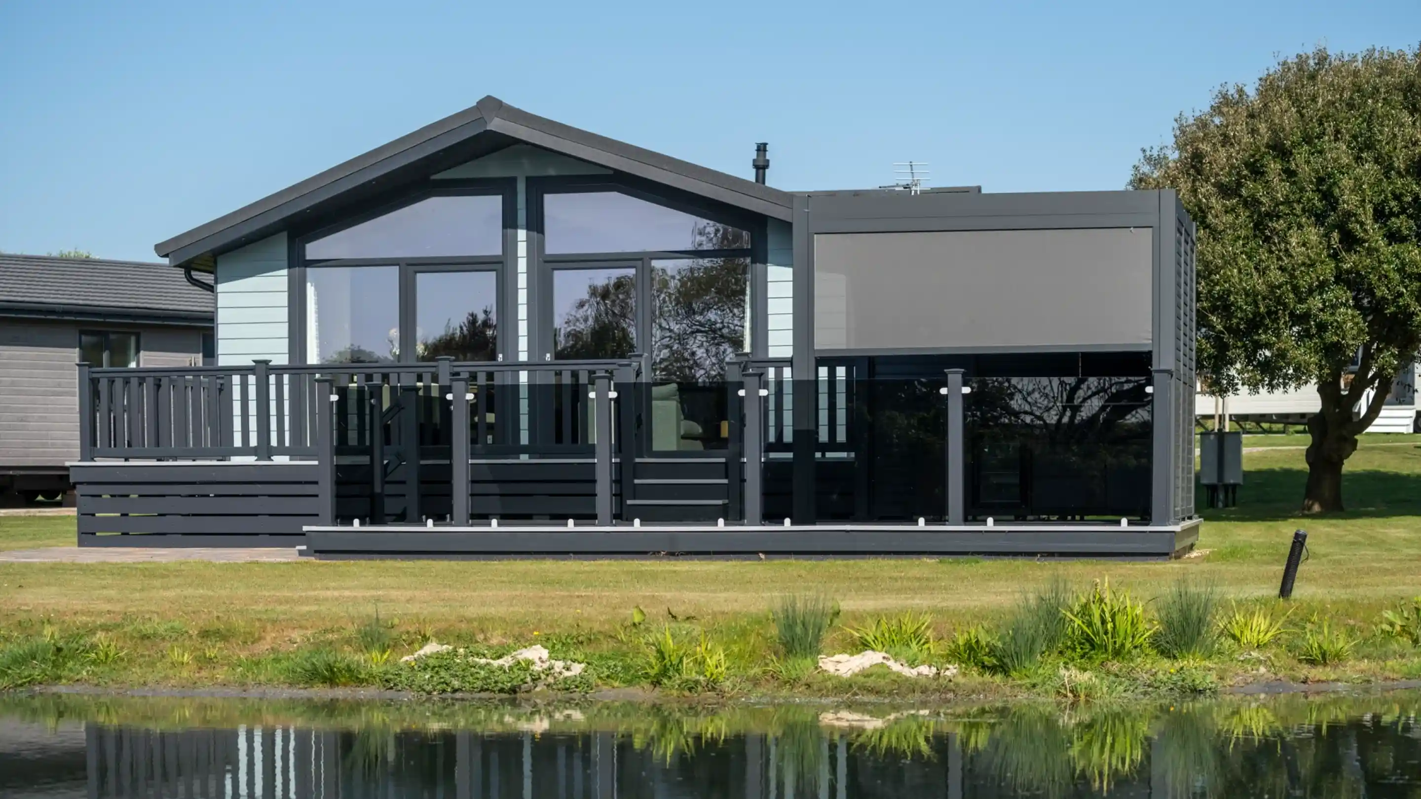 A modern black and gray house with large windows and a deck overlooks a calm pond, surrounded by green grass and shrubs under a clear blue sky.