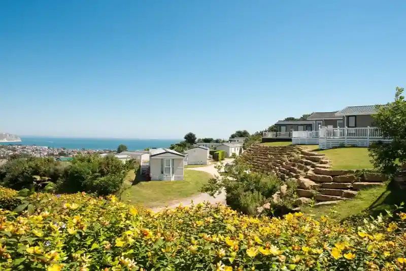 A scenic view of a coastal area featuring several modern cabins on a hillside, surrounded by lush greenery and colorful flowers. The ocean is visible in the background under a clear blue sky.