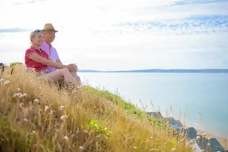 A smiling couple sits together on a grassy hillside overlooking a calm sea. The woman wears a red shirt, while the man dons a white shirt and a straw hat. The sky is clear with a few clouds.