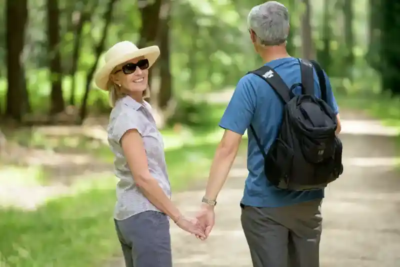 A woman wearing a straw hat and sunglasses holds hands with a man in a blue shirt and backpack as they walk down a tree-lined path. Both are smiling and enjoying their time together.