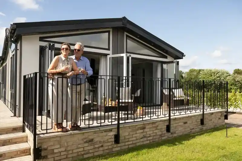 A couple stands on a balcony with a modern home in the background. The man and woman are smiling, dressed casually, with green grass and a landscaped area surrounding the house. The sky is clear and blue.