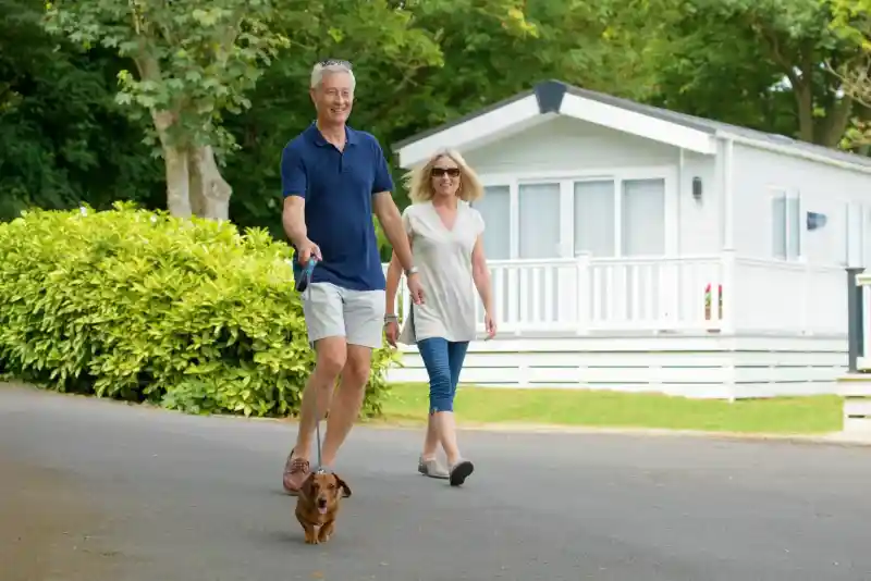 A smiling older man walks a small dog along a path, while a woman casually strolls beside him. They are surrounded by greenery and a white house in the background.