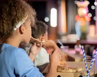 A young girl with curly hair eats a slice of pizza while sitting at a table, watching a performance on stage. A boy is visible beside her, also focused on the show. Colorful stage lights create a festive atmosphere.