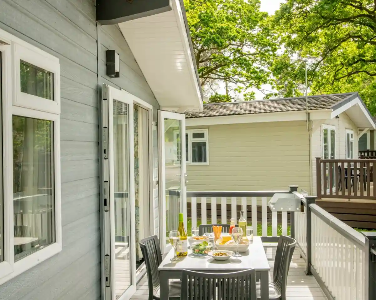 A sunny outdoor patio with a table set for a meal, featuring wine glasses, plates of food, and a view of nearby houses surrounded by lush green trees.