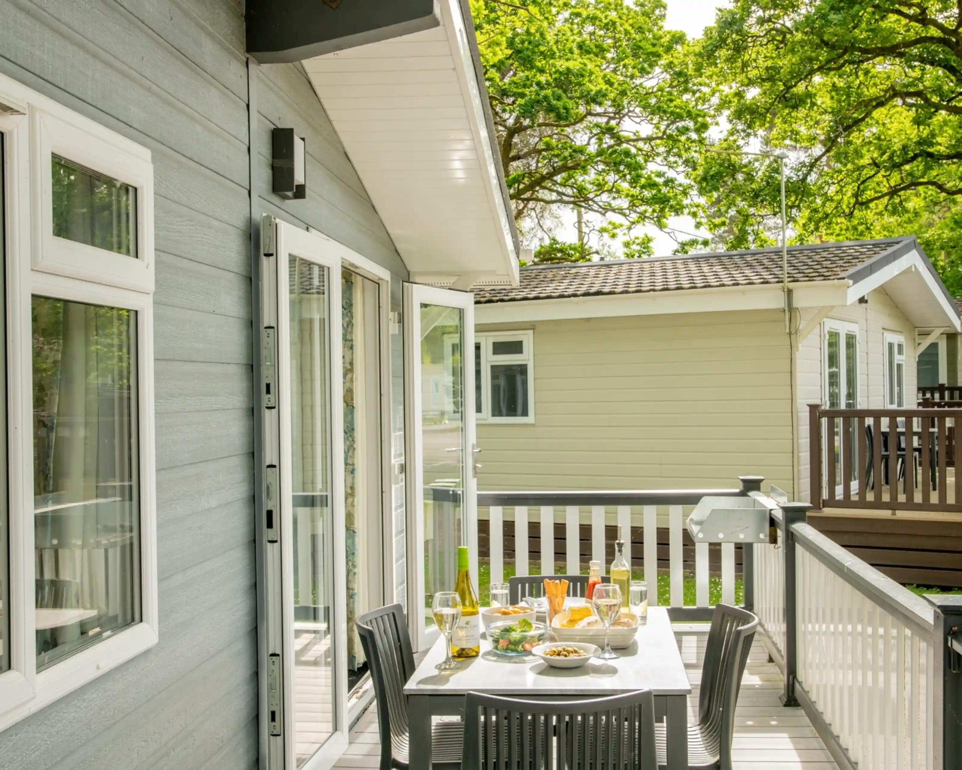 A sunny outdoor patio with a table set for a meal, featuring wine glasses, plates of food, and a view of nearby houses surrounded by lush green trees.
