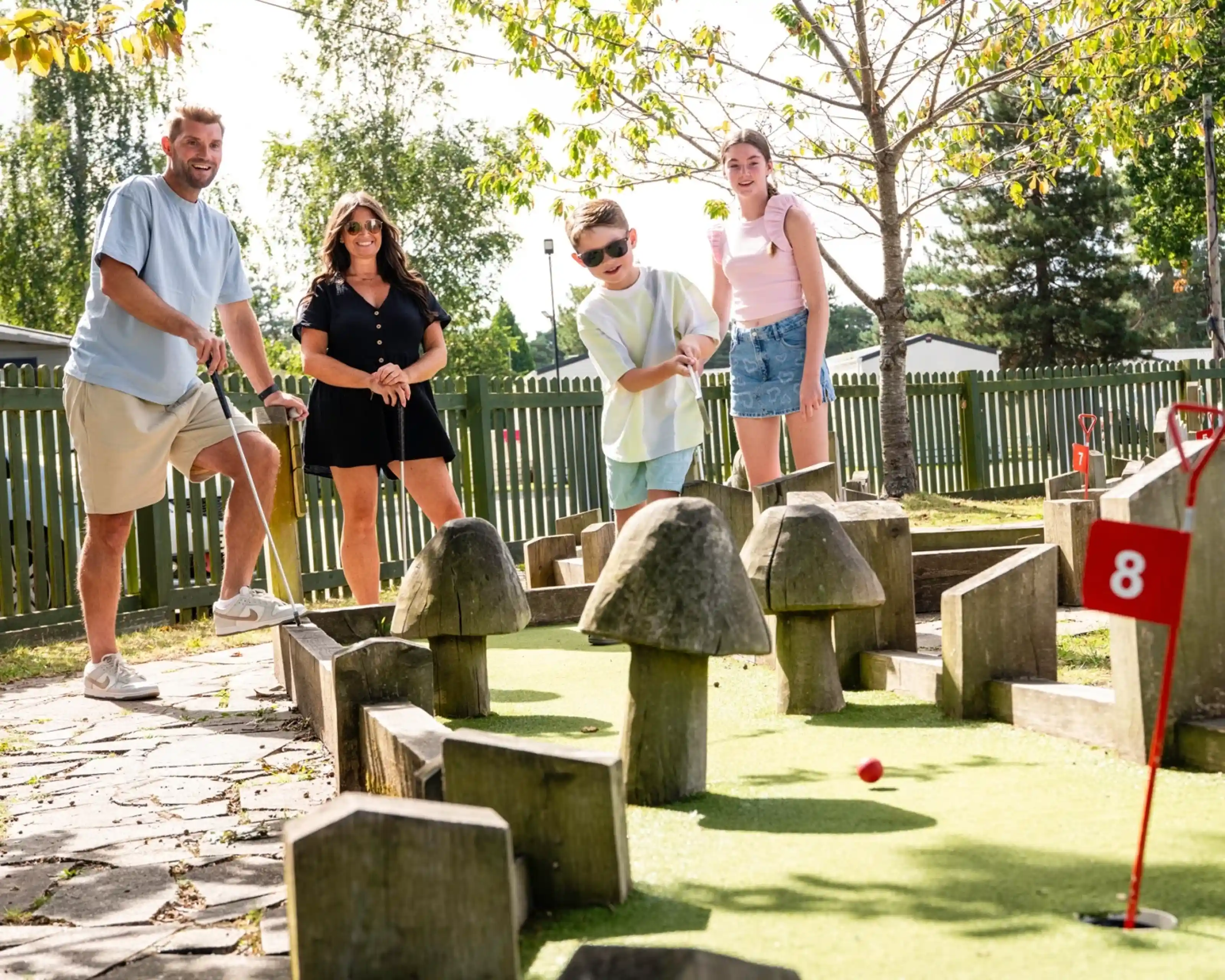 A family enjoys a game of mini-golf on a sunny day. Two adults and two children are engaged, with one child preparing to hit the ball. They are surrounded by whimsical mushroom-shaped obstacles on the green course, with trees and a wooden fence in the background.