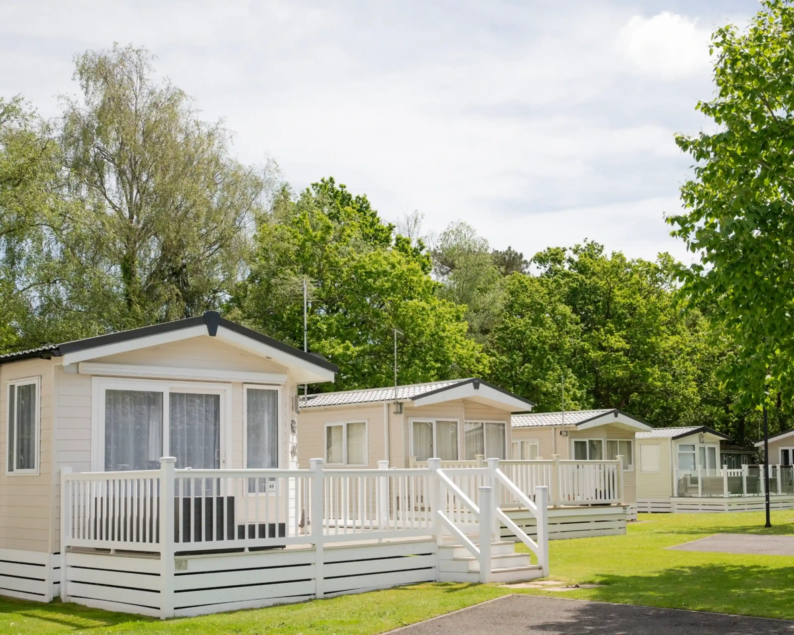 A row of charming white cottages with verandas, surrounded by lush green trees and grass under a bright sky.