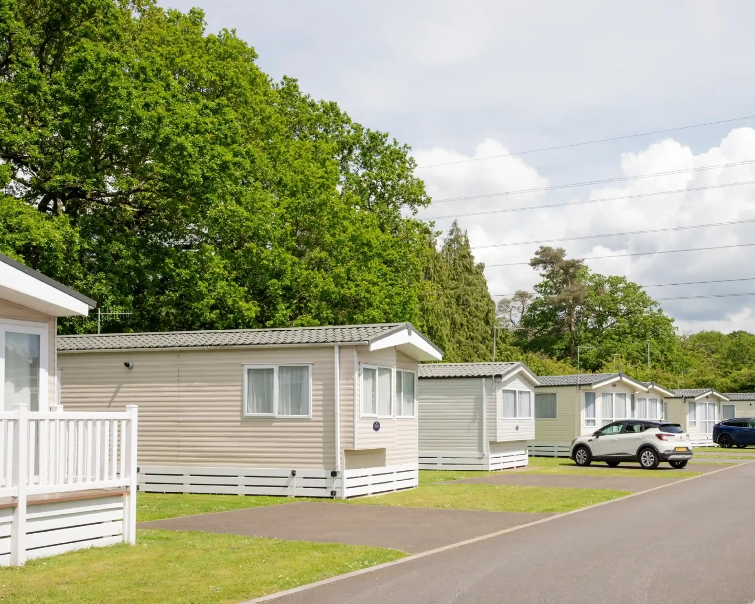 Row of holiday cabins with a neatly paved road in front, surrounded by lush green trees and a partly cloudy sky. A white car is parked in the foreground.