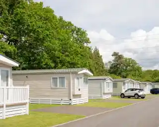 Row of holiday cabins with a neatly paved road in front, surrounded by lush green trees and a partly cloudy sky. A white car is parked in the foreground.