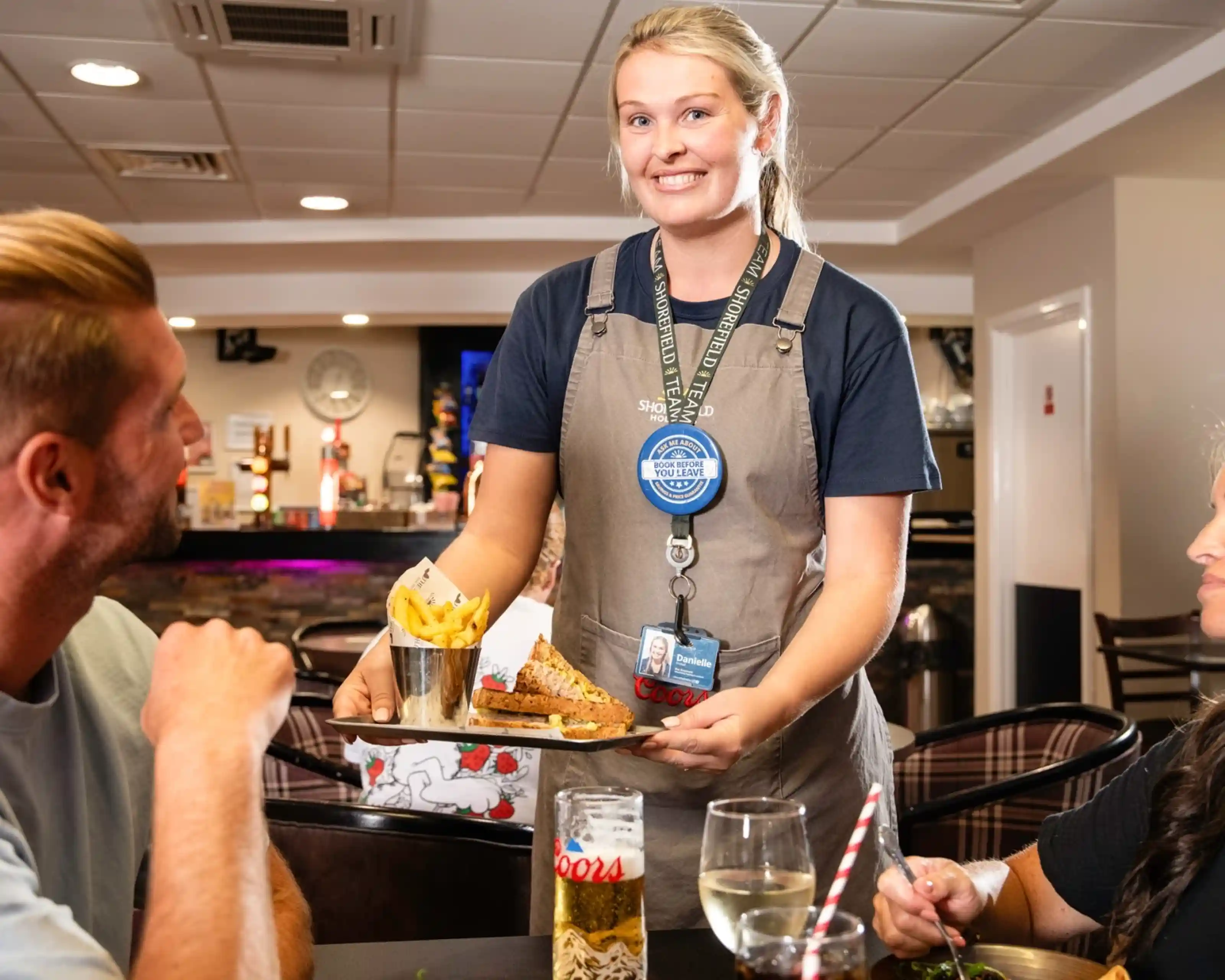 A waitress smiles while serving a plate of food and fries to two seated customers in a casual dining setting. A can of Coors beer is visible on the table.