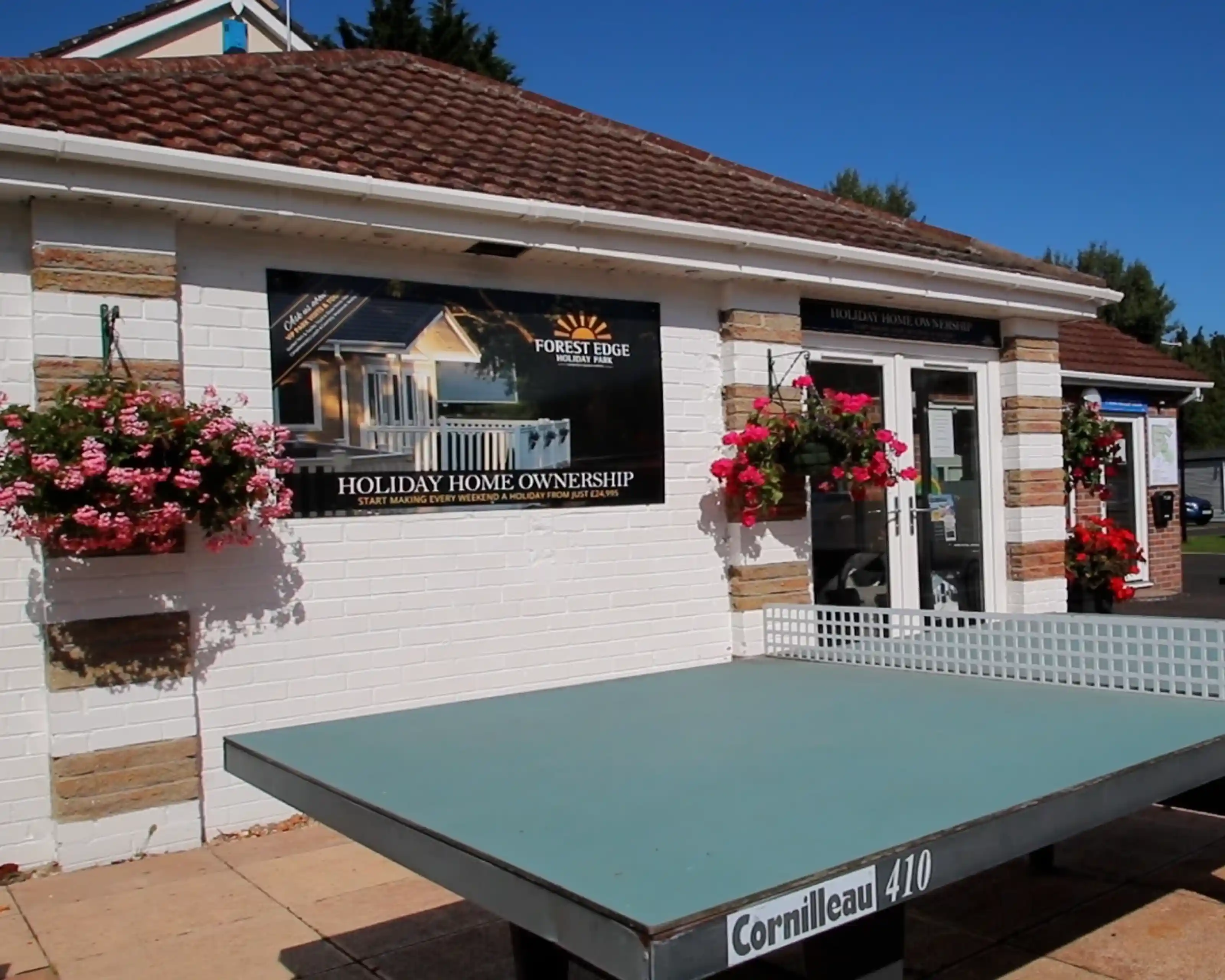 A ping pong table with a sign promoting holiday home ownership outside a building. The building features hanging flower baskets and a welcoming entrance.