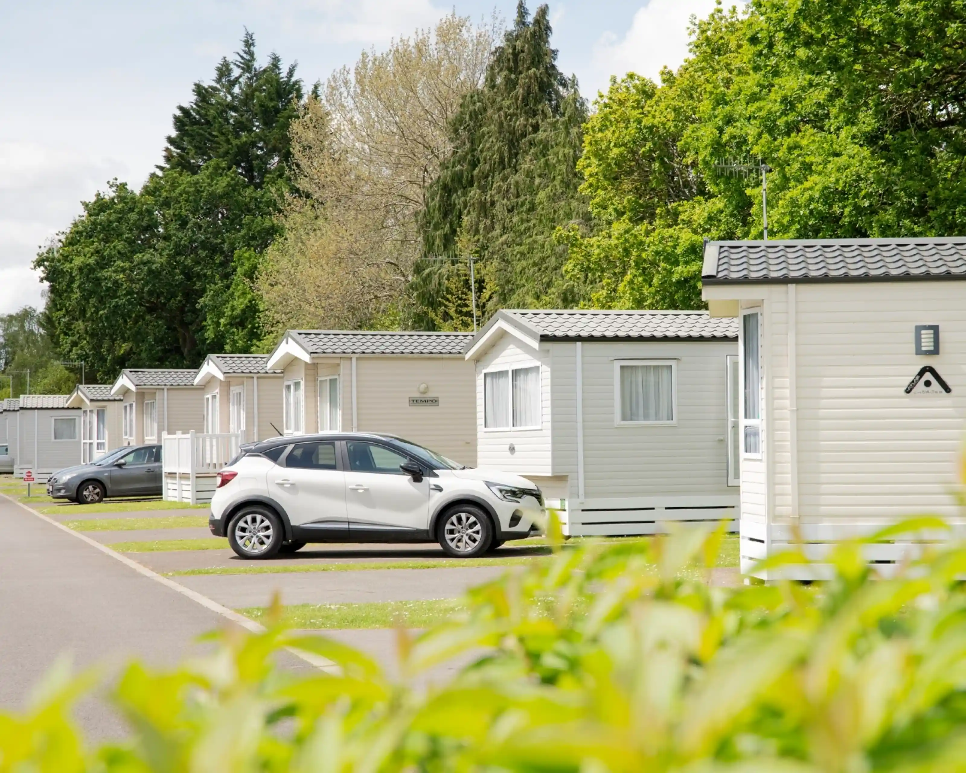 Row of holiday cabins lined up along a paved pathway, with a white car parked in front of one cabin. Lush greenery and trees provide a backdrop.