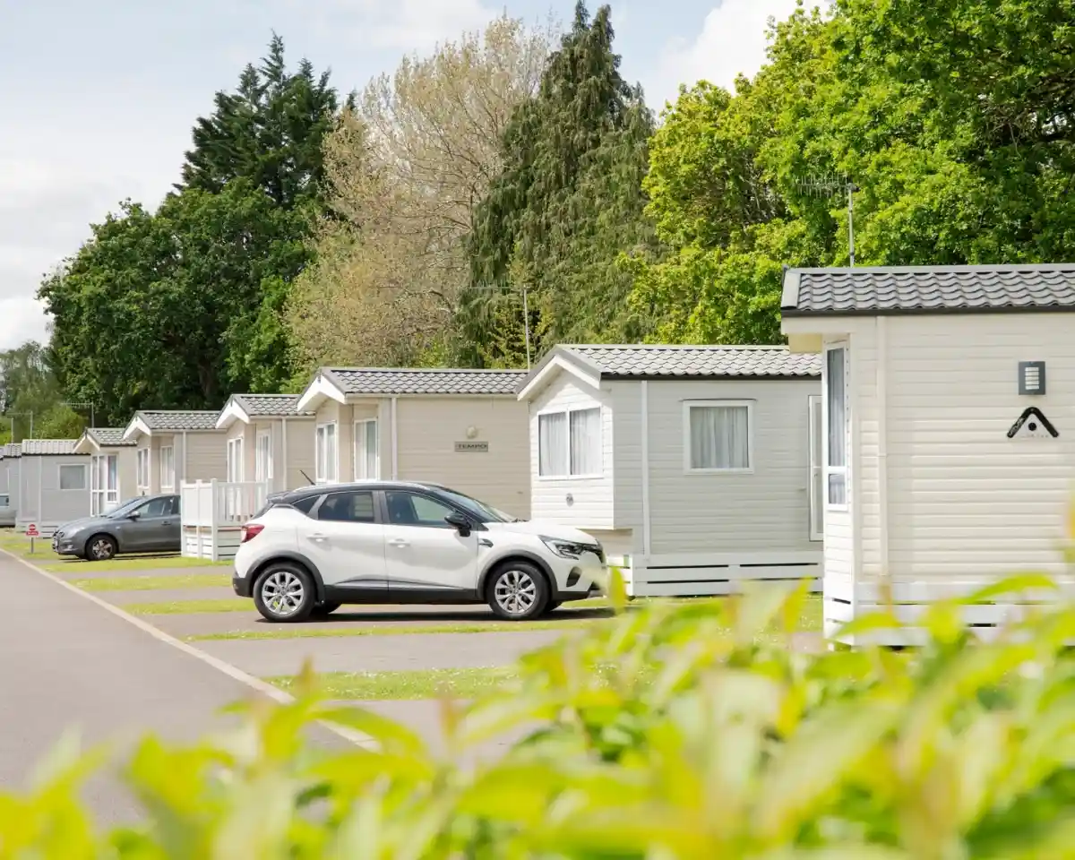 Row of holiday cabins lined up along a paved pathway, with a white car parked in front of one cabin. Lush greenery and trees provide a backdrop.