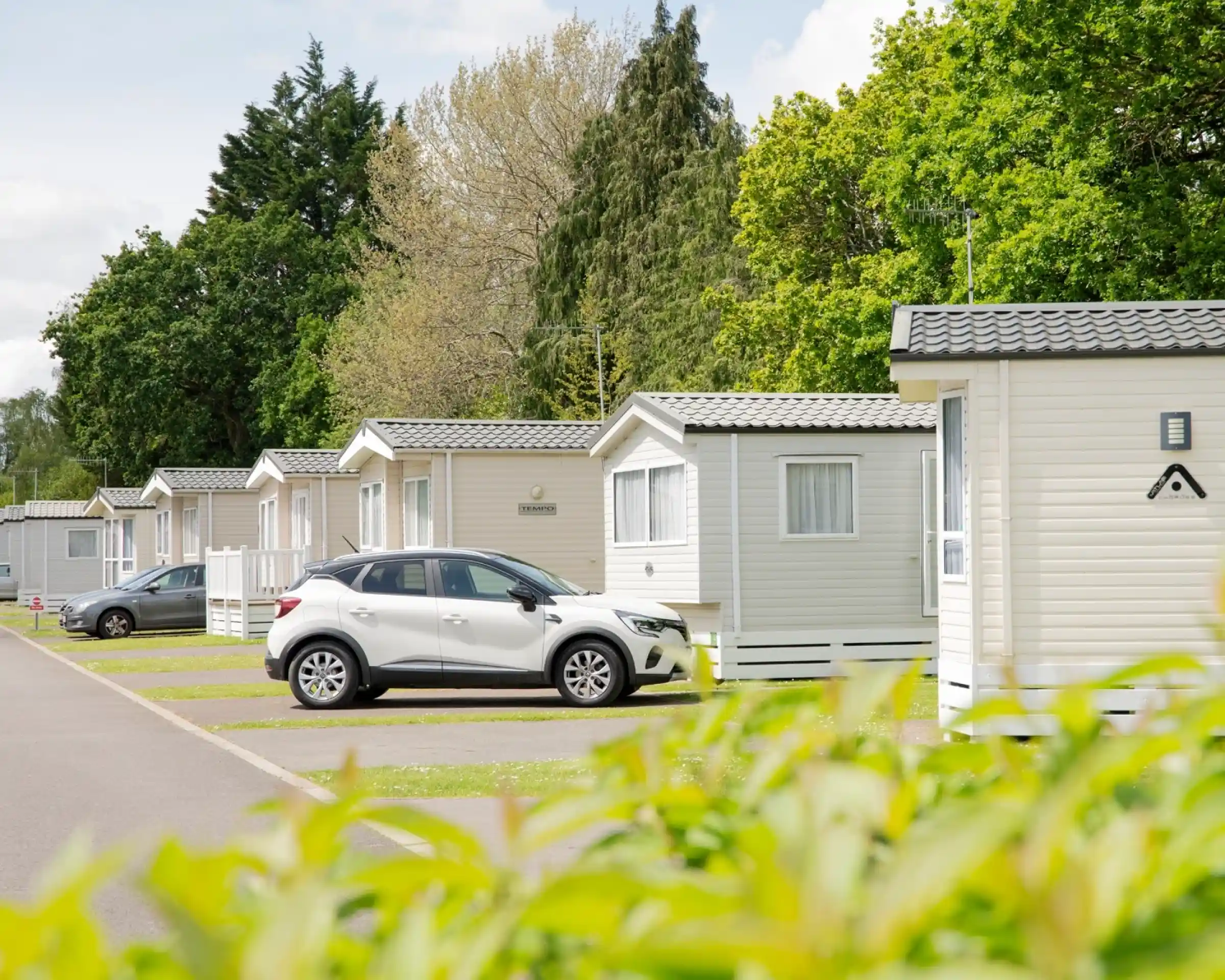 Row of holiday cabins lined up along a paved pathway, with a white car parked in front of one cabin. Lush greenery and trees provide a backdrop.