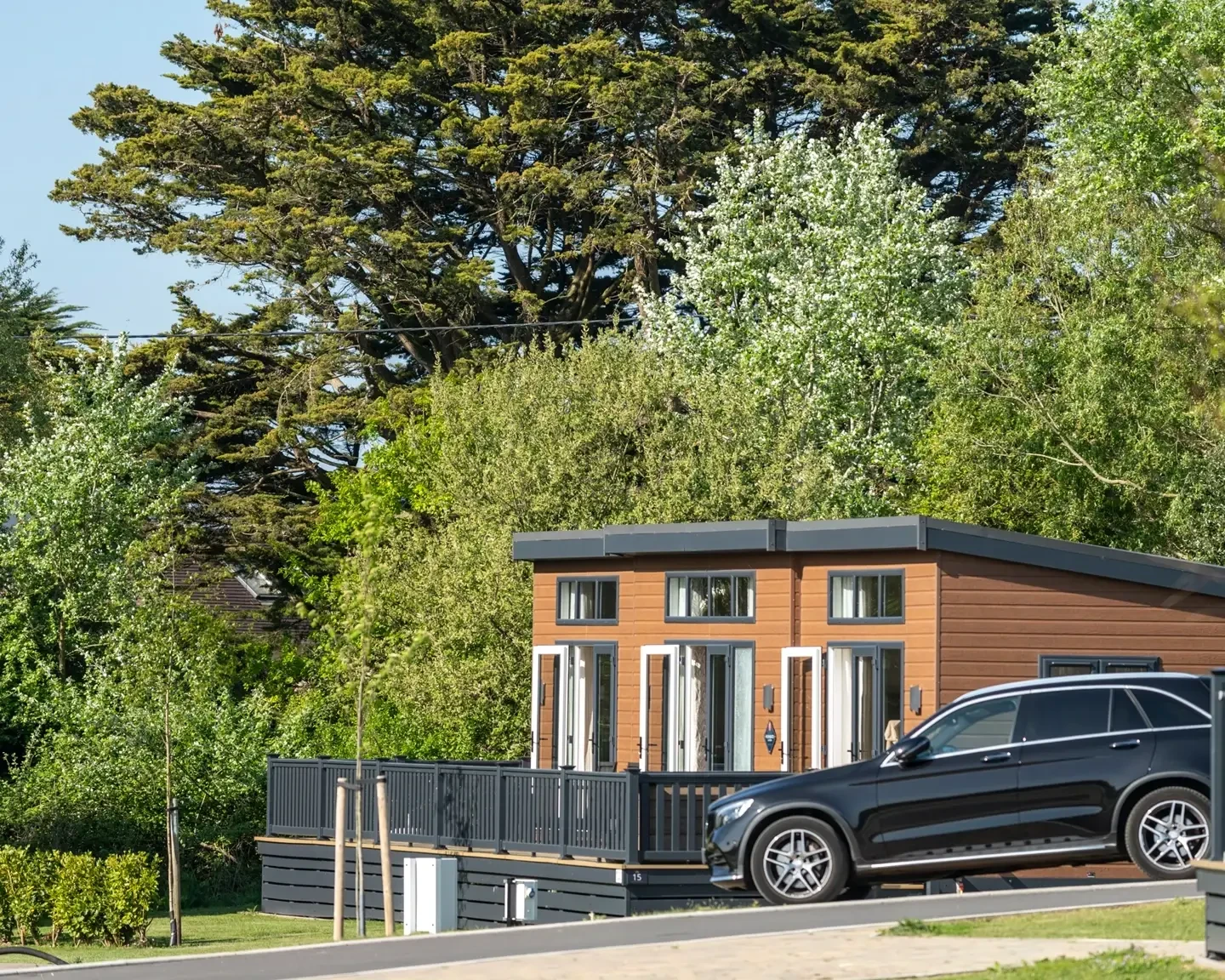 A modern wooden cabin with a dark exterior and large windows, surrounded by lush greenery. The cabin features a spacious deck with a railing, and a black car is parked near the cabin. Tall trees provide shade in the background, and the scene is illuminated by bright sunlight, highlighting the peaceful, natural environment.