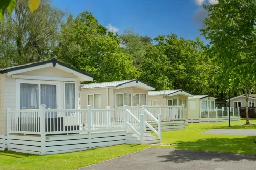 Charming holiday cottages with white railings set in a green, sunny park. Lush trees provide shade, and a clear blue sky adds brightness to the scene.