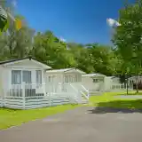 Charming holiday cottages with white railings set in a green, sunny park. Lush trees provide shade, and a clear blue sky adds brightness to the scene.