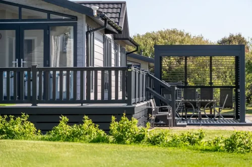 A modern wooden house with large windows and a dark exterior, featuring a spacious deck. In the foreground, a manicured lawn leads to a stylish outdoor seating area with a table and chairs under a covered pavilion. Lush trees line the background under a clear blue sky.
