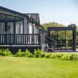 A modern wooden house with large windows and a dark exterior, featuring a spacious deck. In the foreground, a manicured lawn leads to a stylish outdoor seating area with a table and chairs under a covered pavilion. Lush trees line the background under a clear blue sky.