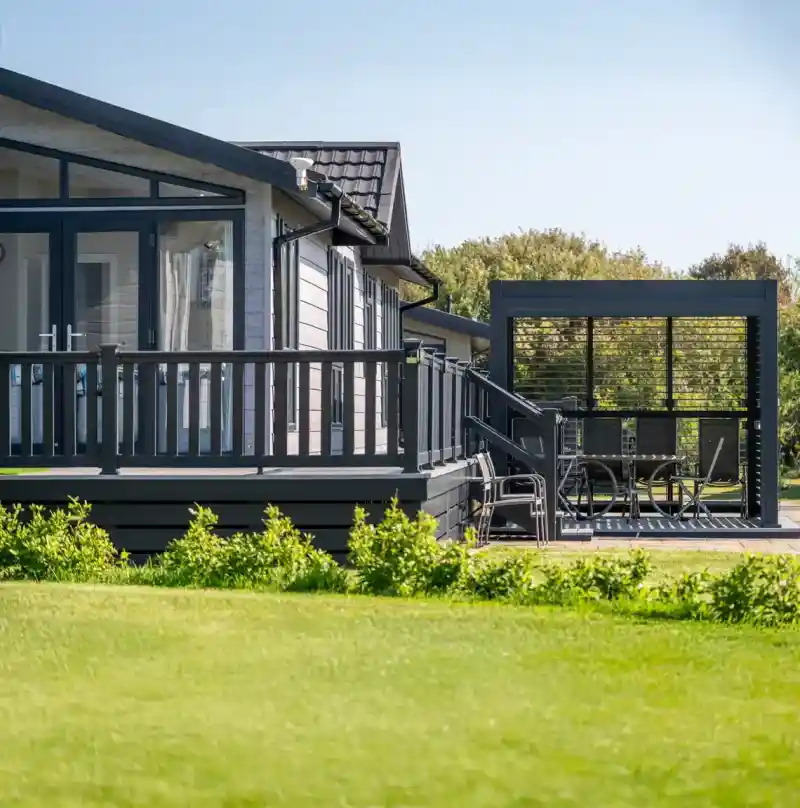 A modern wooden house with large windows and a dark exterior, featuring a spacious deck. In the foreground, a manicured lawn leads to a stylish outdoor seating area with a table and chairs under a covered pavilion. Lush trees line the background under a clear blue sky.