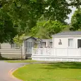 Two mobile homes are set beside a well-maintained green lawn, surrounded by leafy trees. A white deck with an umbrella is visible in front of one home.