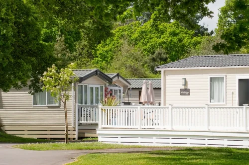 Two mobile homes are set beside a well-maintained green lawn, surrounded by leafy trees. A white deck with an umbrella is visible in front of one home.
