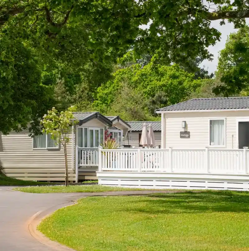Two mobile homes are set beside a well-maintained green lawn, surrounded by leafy trees. A white deck with an umbrella is visible in front of one home.