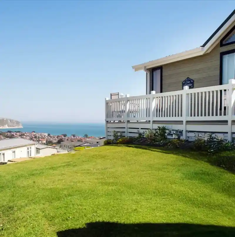 A well-maintained grassy yard in the foreground, leading up to a house with a white picket fence. In the background, the ocean stretches out, with a coastal town visible along the shore under a clear blue sky.
