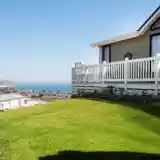 A well-maintained grassy yard in the foreground, leading up to a house with a white picket fence. In the background, the ocean stretches out, with a coastal town visible along the shore under a clear blue sky.