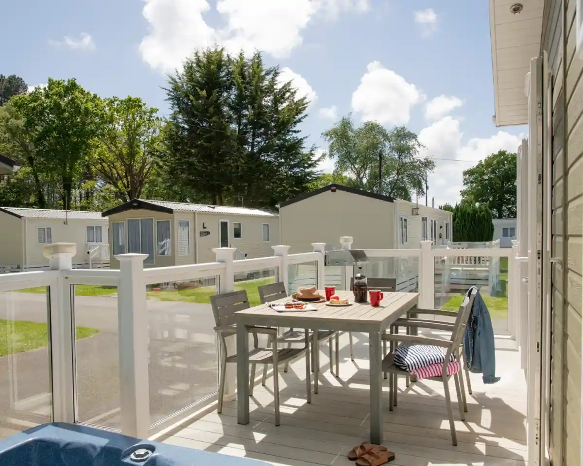 A sunny outdoor patio with a wooden dining table set for four, featuring red mugs and a plate of snacks. Nearby, a blue towel hangs on a railing. In the background, holiday homes are visible, surrounded by greenery and a clear blue sky.