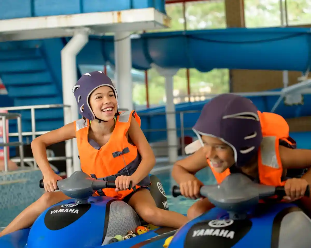 Two children wearing orange life vests and helmets are joyfully riding jet skis in a pool. They are smiling and focused, with a blue waterslide and pool structure visible in the background.