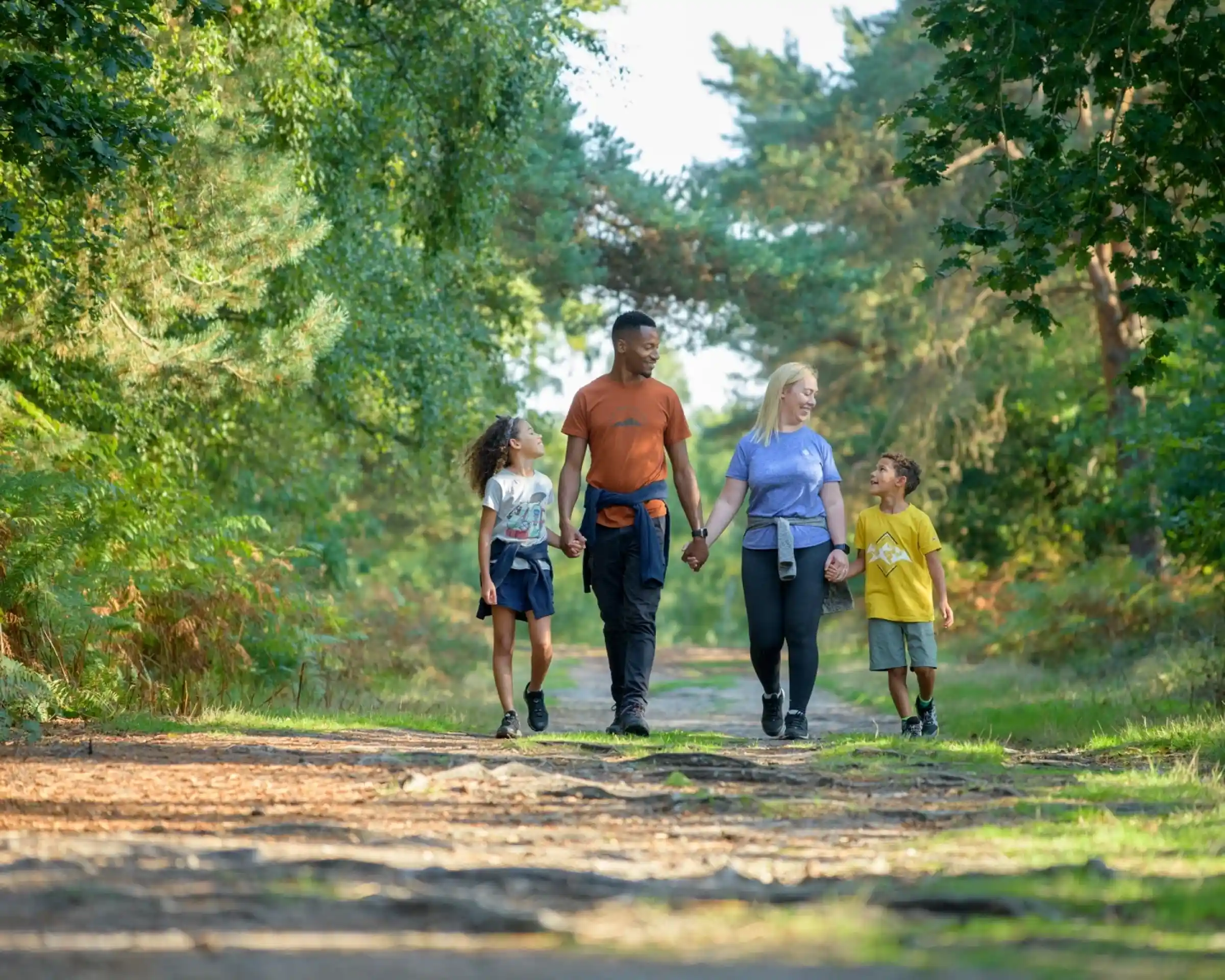 A family of four walks hand in hand along a trail in a forest, surrounded by green trees and plants. Two children, a boy and a girl, are on the left, while their parents, a man and a woman, walk beside them. The setting is bright and sunny.