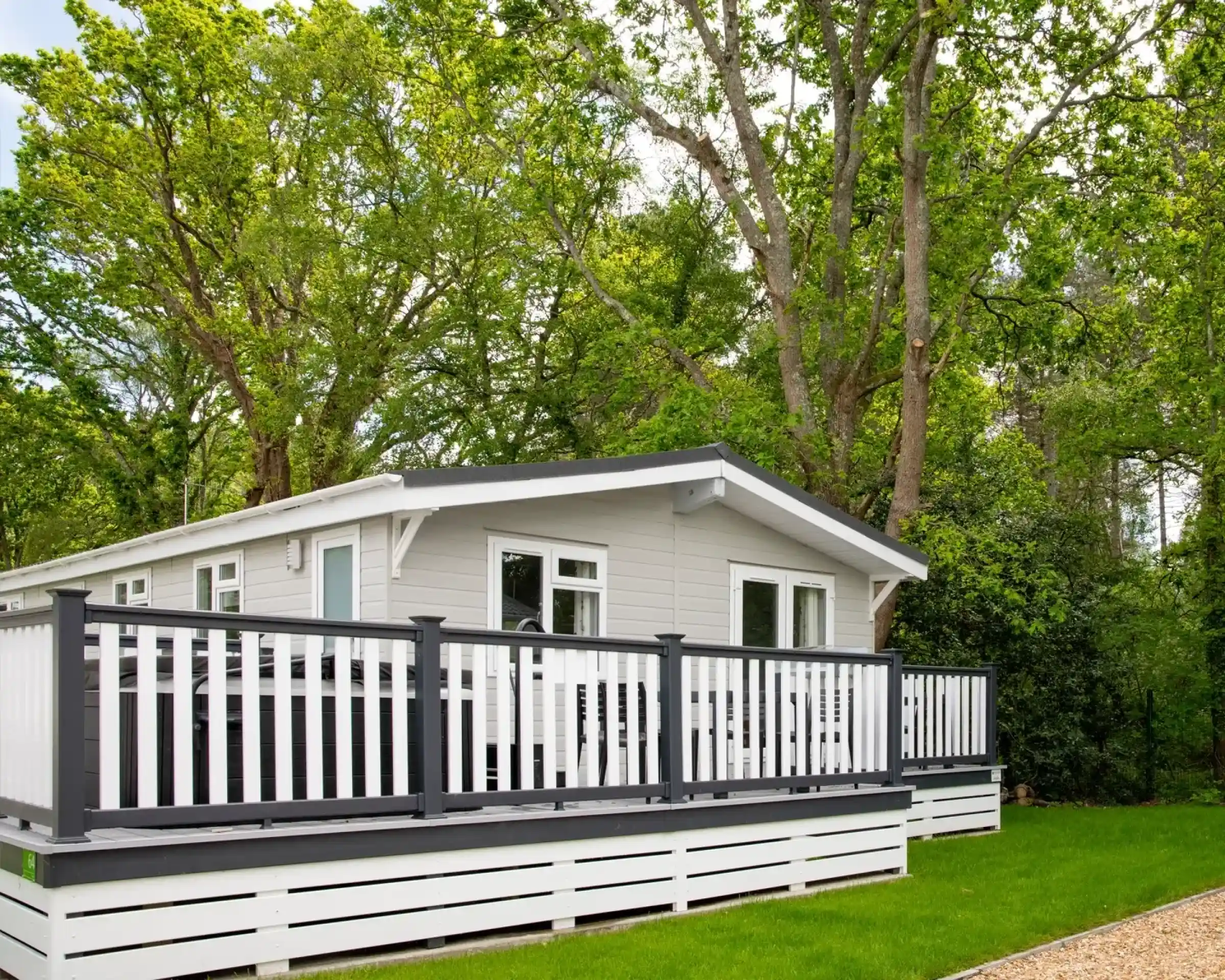 A modern cabin with a gray and white exterior, featuring a spacious deck with black railings. Surrounding it are lush green trees, indicating a serene, wooded environment.