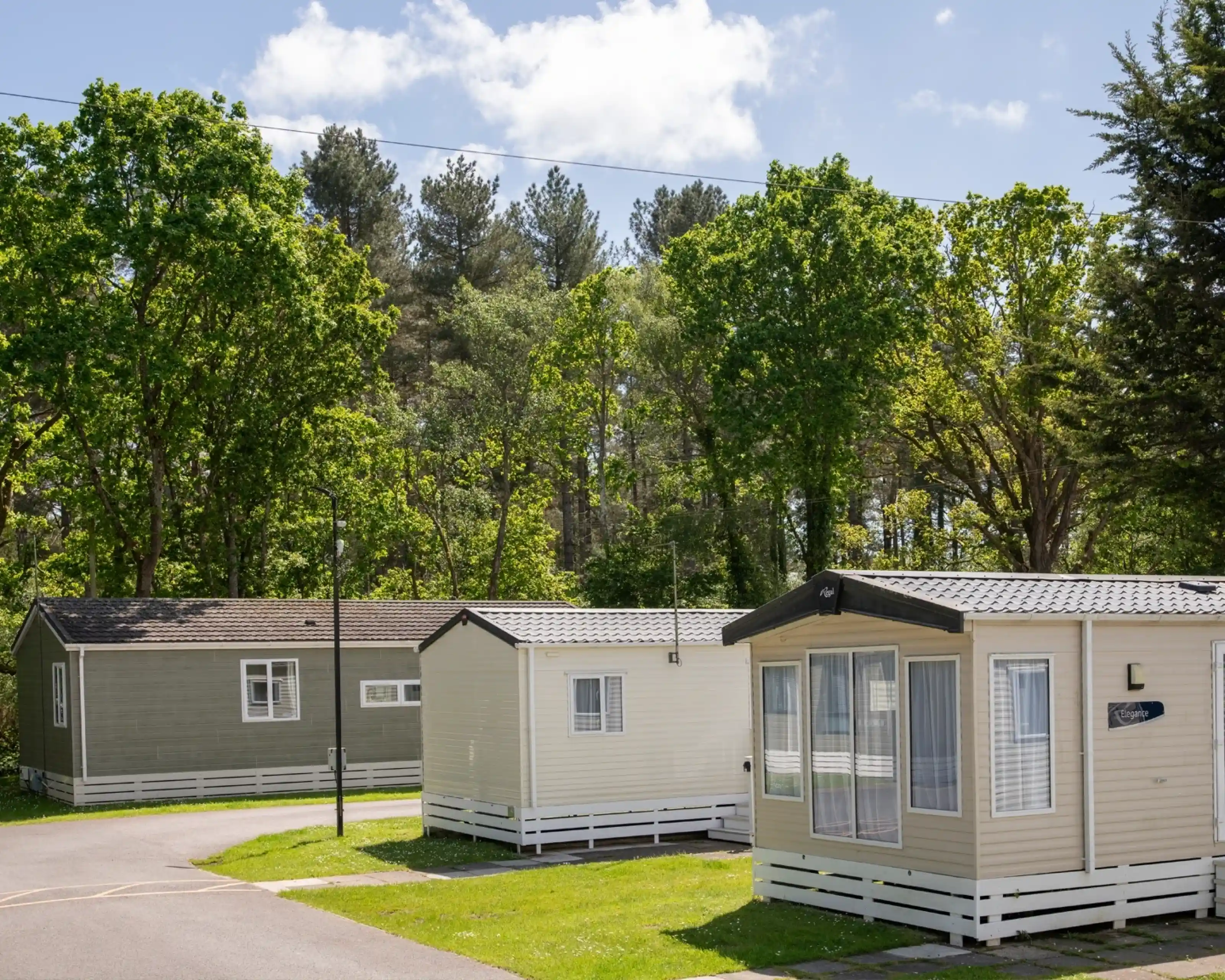 A row of static caravans in a lush green campsite surrounded by trees under a blue sky with scattered clouds.