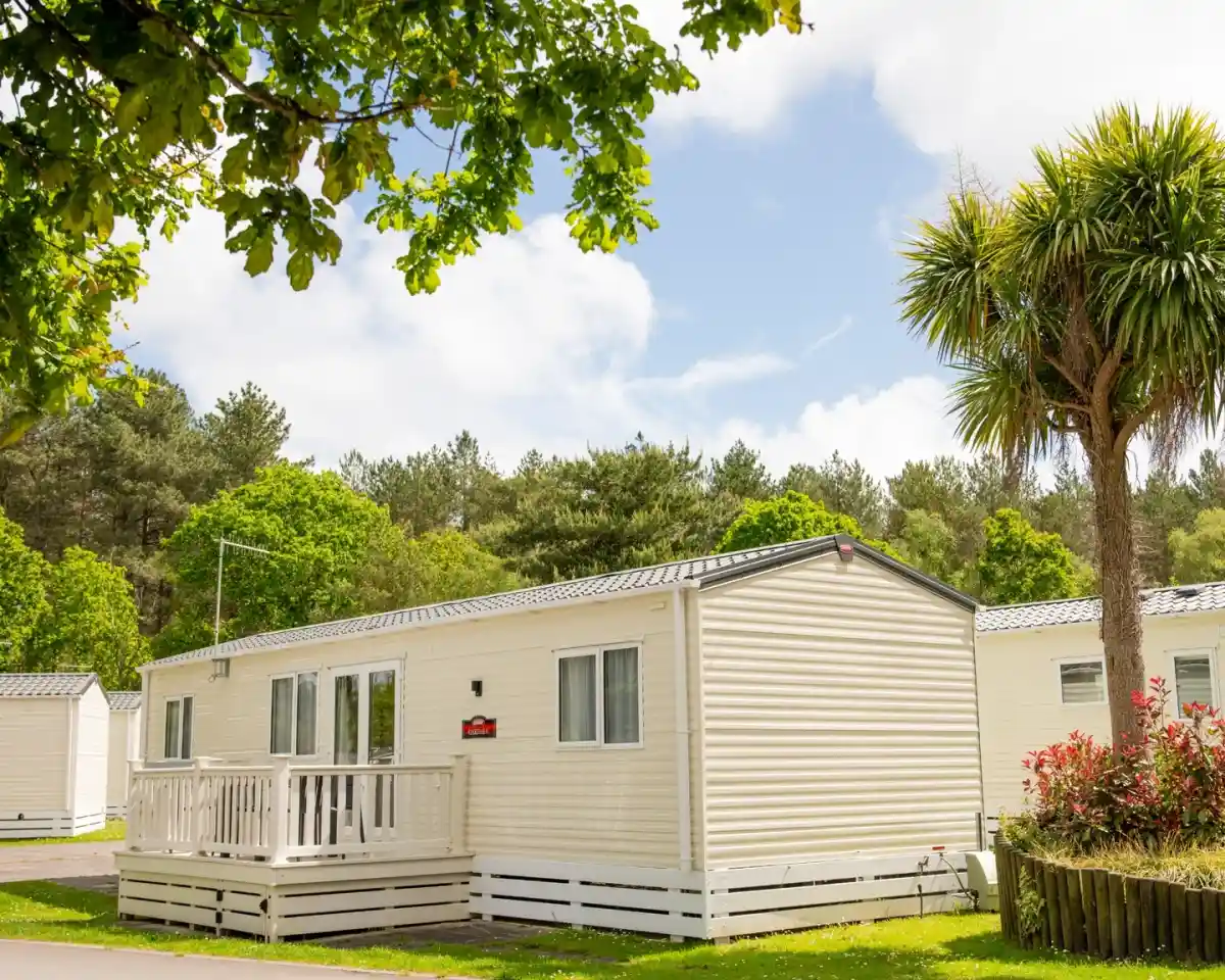 A light-colored holiday home with a small porch, surrounded by lush green trees and plants. The sky is partly cloudy, creating a bright, scenic atmosphere.