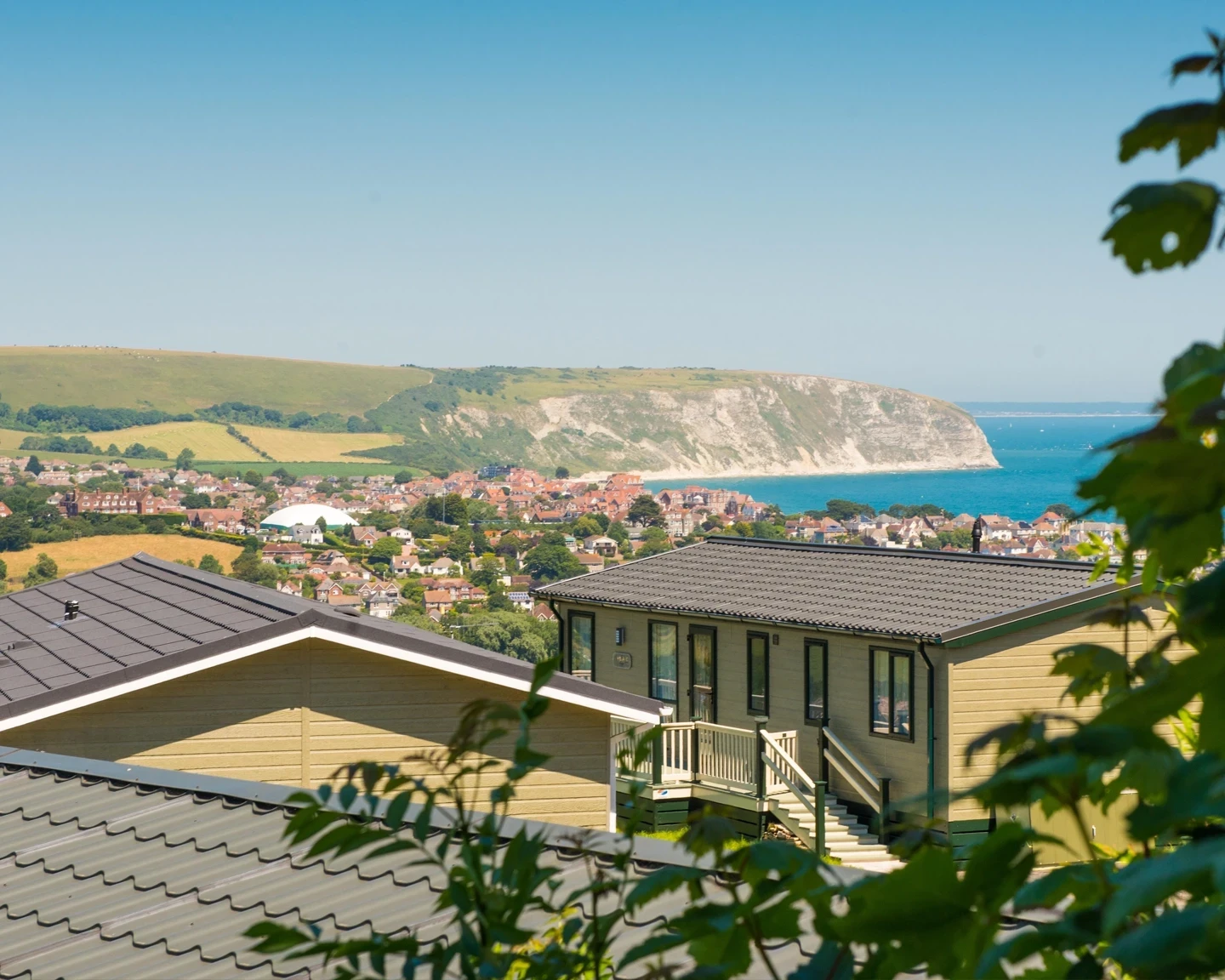 A scenic view of a coastal village with colorful houses and green hills, framed by rooftops in the foreground. The ocean glimmers under a clear blue sky, with cliffs visible in the distance.