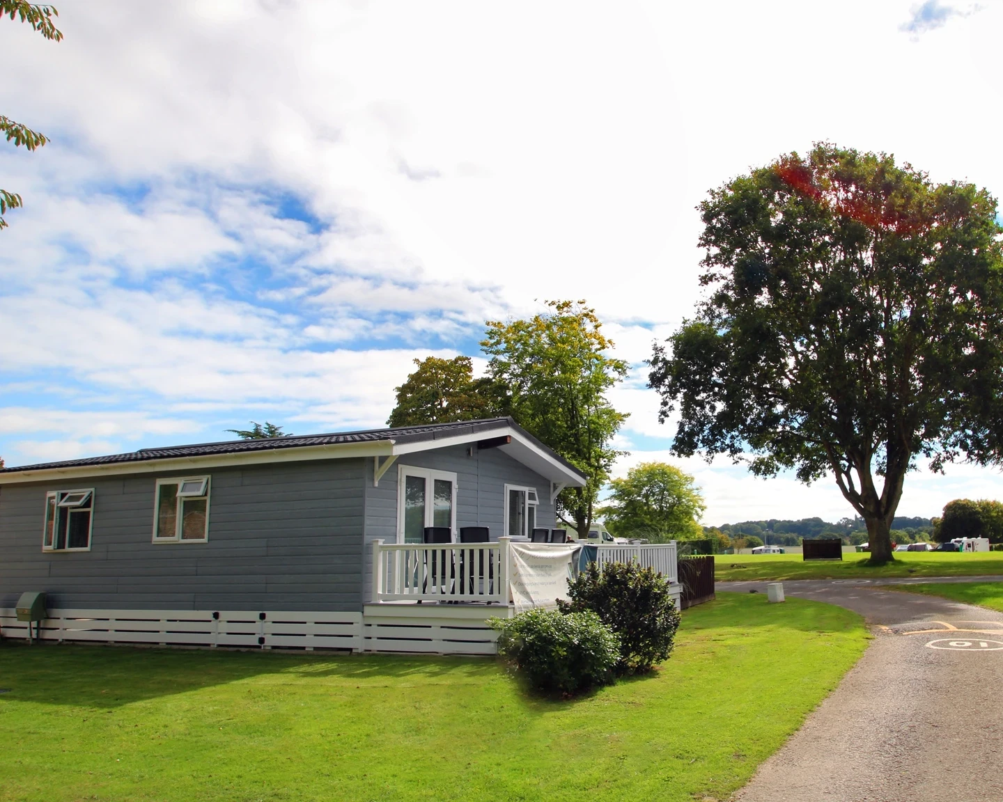 A grey single-story mobile home with a white porch sits on a grassy area, surrounded by trees. A winding pathway leads up to the home, with a blue sky and fluffy clouds in the background.