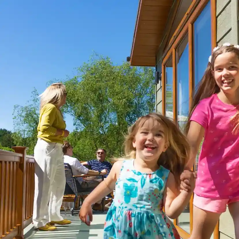 Two young girls hold hands and run joyfully on a sunny deck. One girl wears a blue dress while the other is in a pink outfit. In the background, adults are seated and engaged in conversation. Bright blue sky and greenery enhance the cheerful atmosphere.