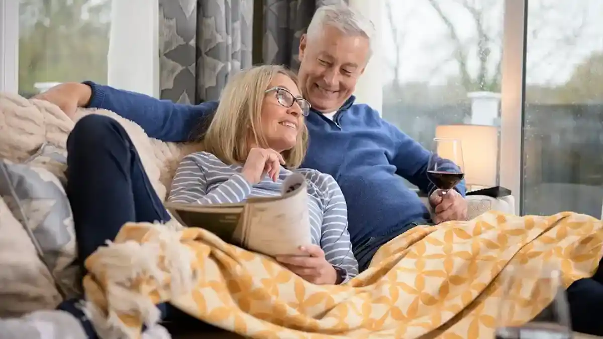 An older couple relaxes on a couch, sharing a cosy moment together. One partner reads a book, while the other smiles and holds a glass of red wine. A warm blanket is draped over them, and a lamp provides soft lighting in the background.