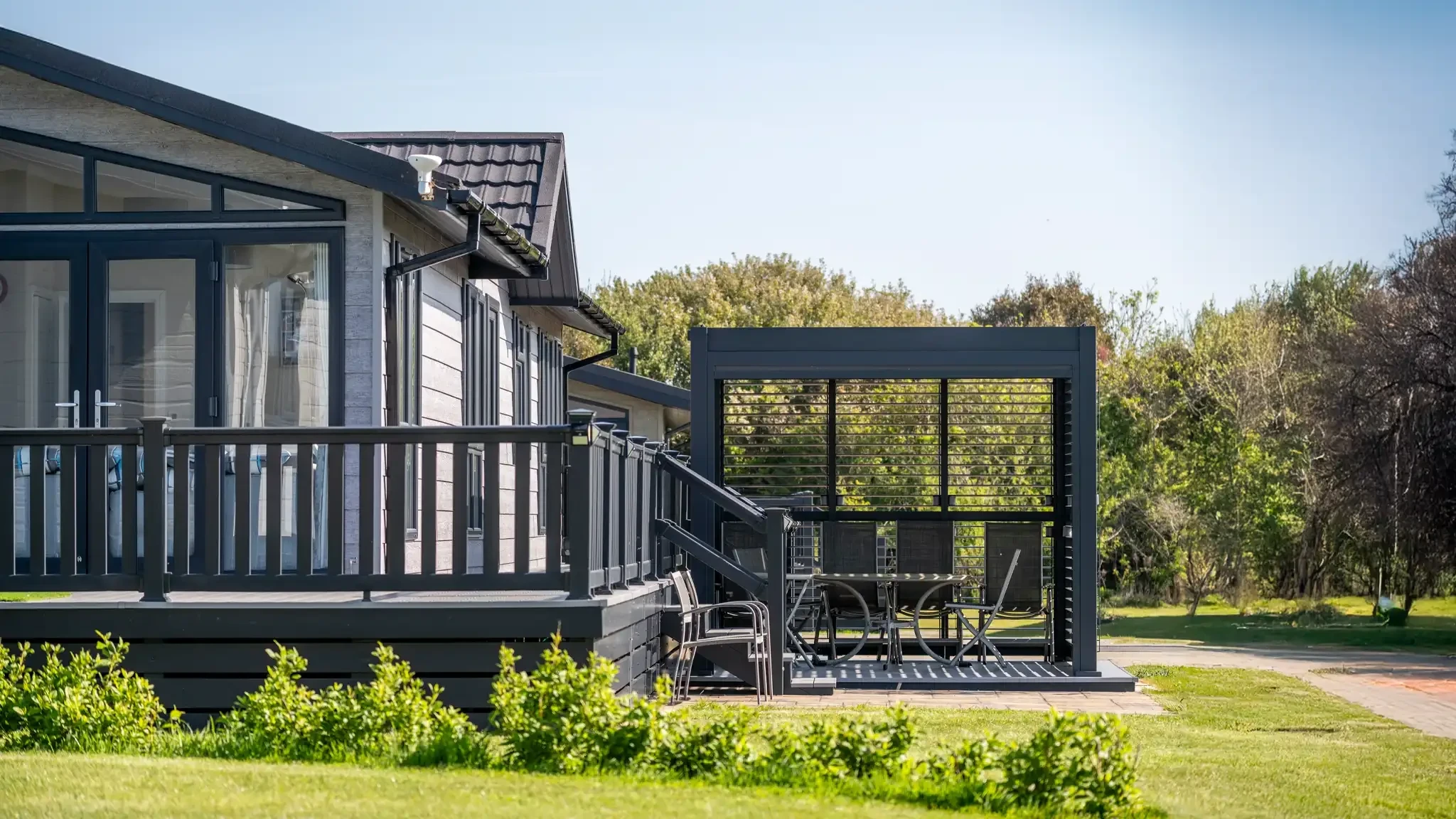 A modern lodge with a rustic design featuring a dark deck and large windows, surrounded by greenery. A covered outdoor seating area with a table and chairs is visible.