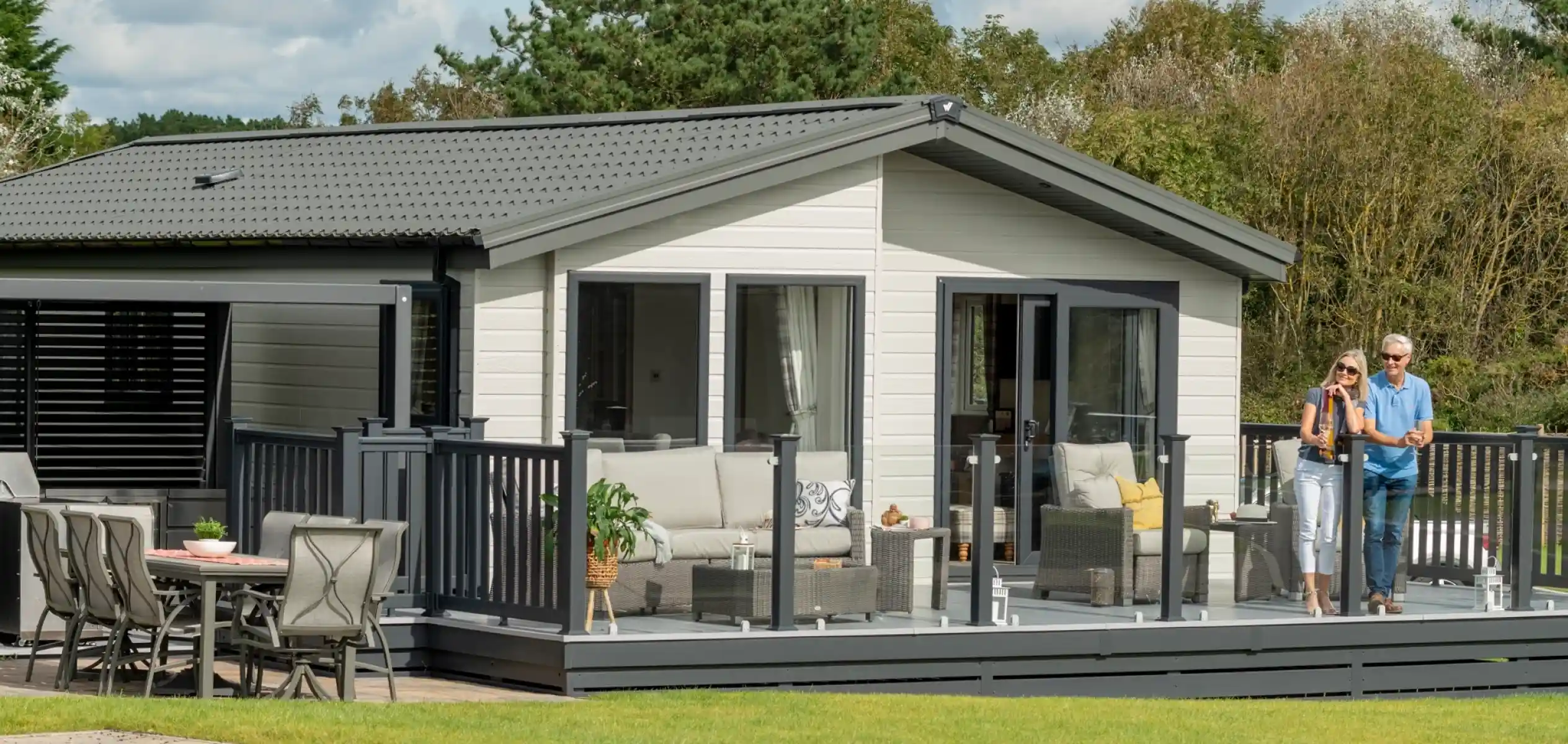A modern lodge holiday home with a spacious deck featuring comfortable seating. Two people stand on the deck, enjoying the outdoor space. Lush greenery surrounds the home, and the sky is partly cloudy.
