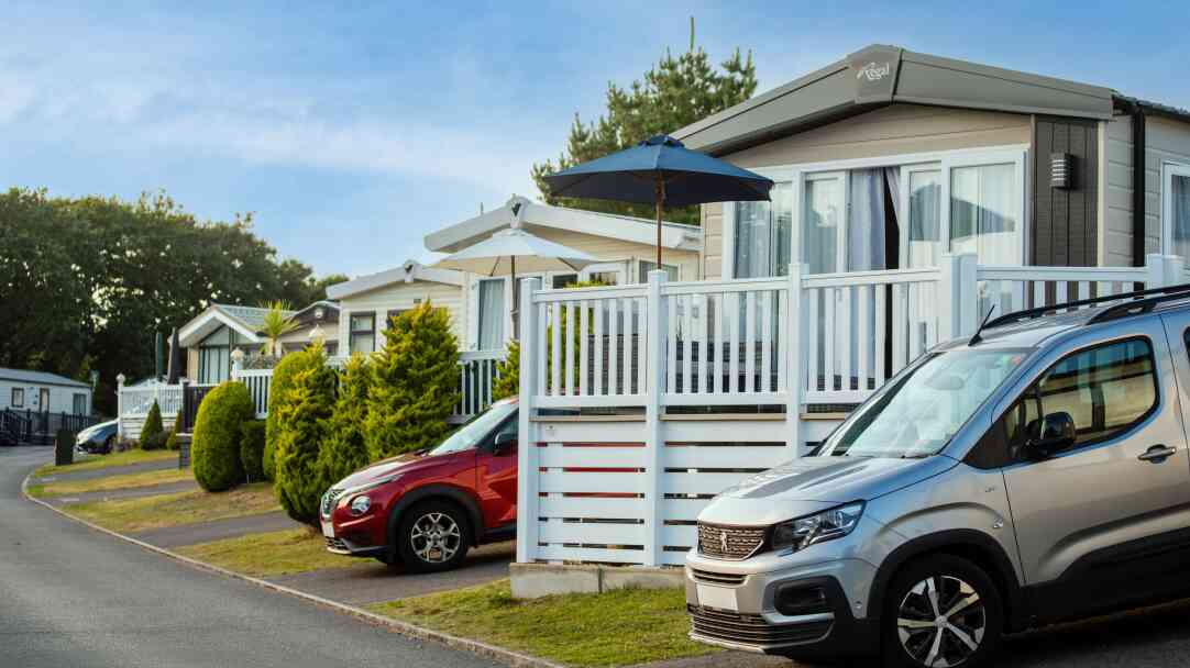 Colorful holiday caravans lined along a paved road, with an umbrella-shaded deck in front of one. A silver vehicle is parked beside a red car, with manicured hedges lining the pathway. Bright blue sky overhead.