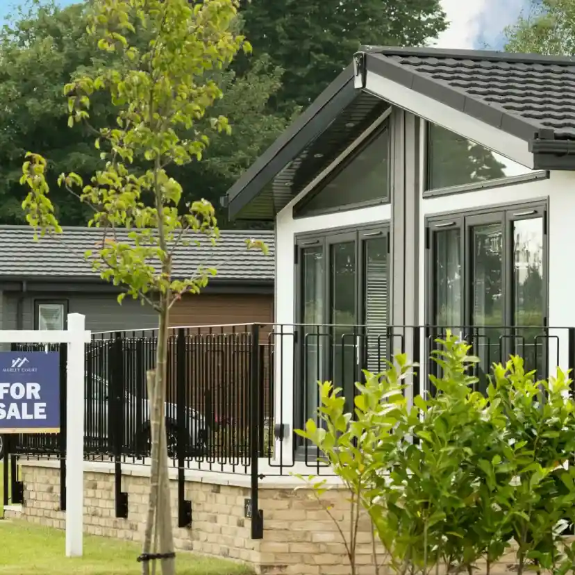 A modern house with large windows and a black roof is shown. In front, a "For Sale" sign stands beside a landscaped area with small bushes. Nearby, several similar houses are visible along a well-maintained pathway.