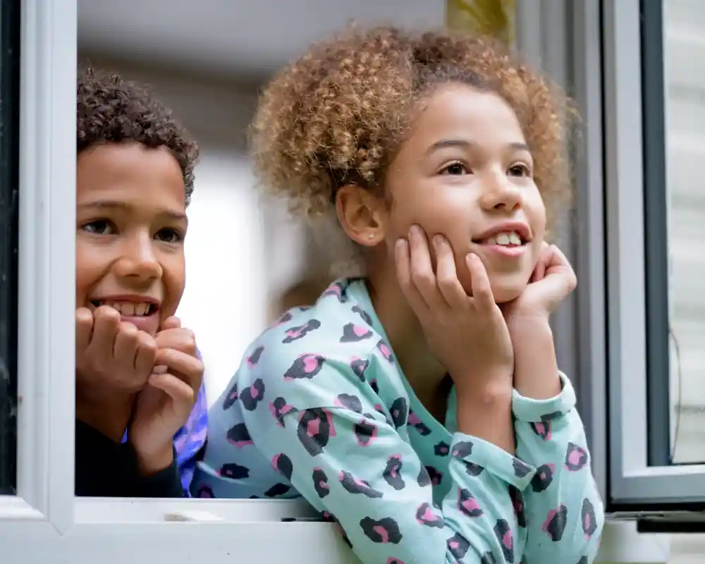 Two children with curly hair peering out of a window, looking excited. One child has dark hair and a light blue shirt with dark spots, while the other has light brown hair and wears a black shirt. They both have their hands on their cheeks, showing curious expressions.