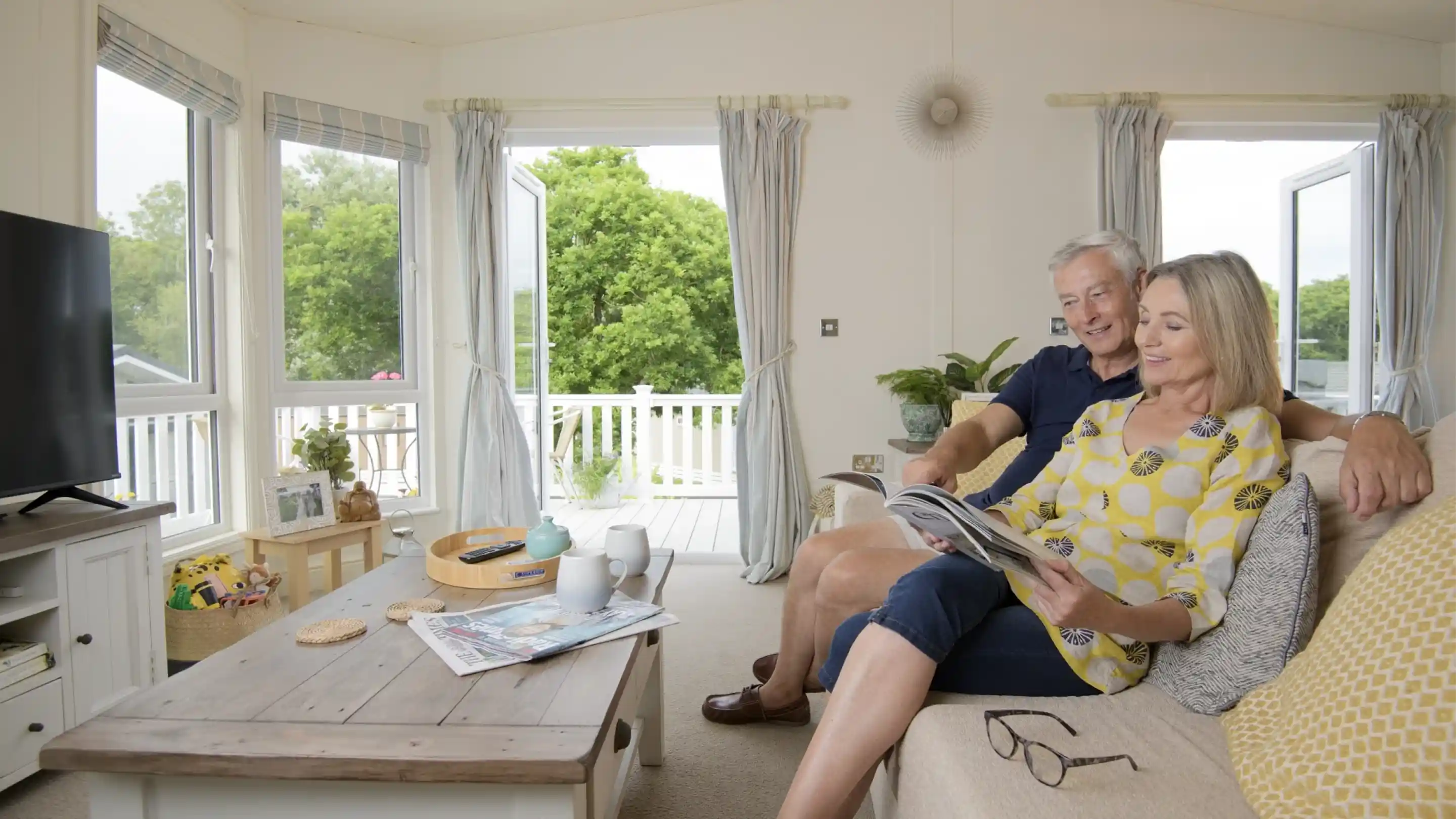 A couple sits together on a couch in a bright, cozy living room, reading a magazine and smiling at each other. Sunlight streams through large windows, illuminating plants and decorative elements.