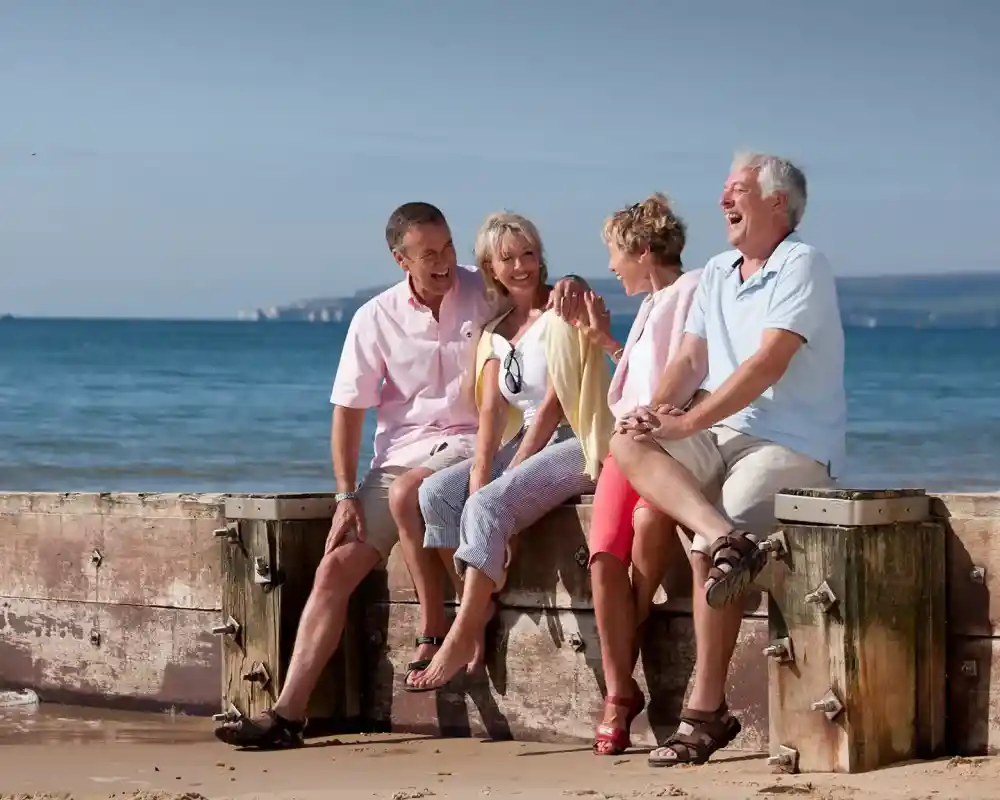 Four adults sit on a wooden barrier at the beach, smiling and laughing together. The sea and a clear sky serve as a backdrop, creating a joyful and relaxed atmosphere.