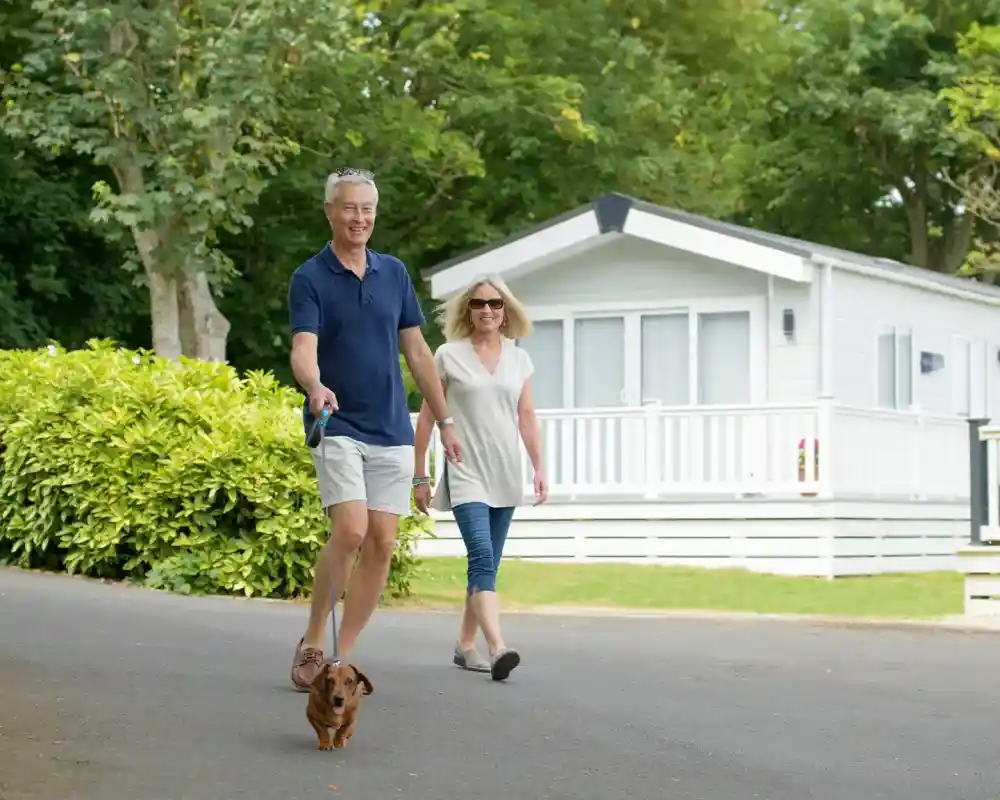 A man and a woman walk along a tree-lined path, smiling as they stroll with a small dog. In the background, a white vacation home with a porch is visible.