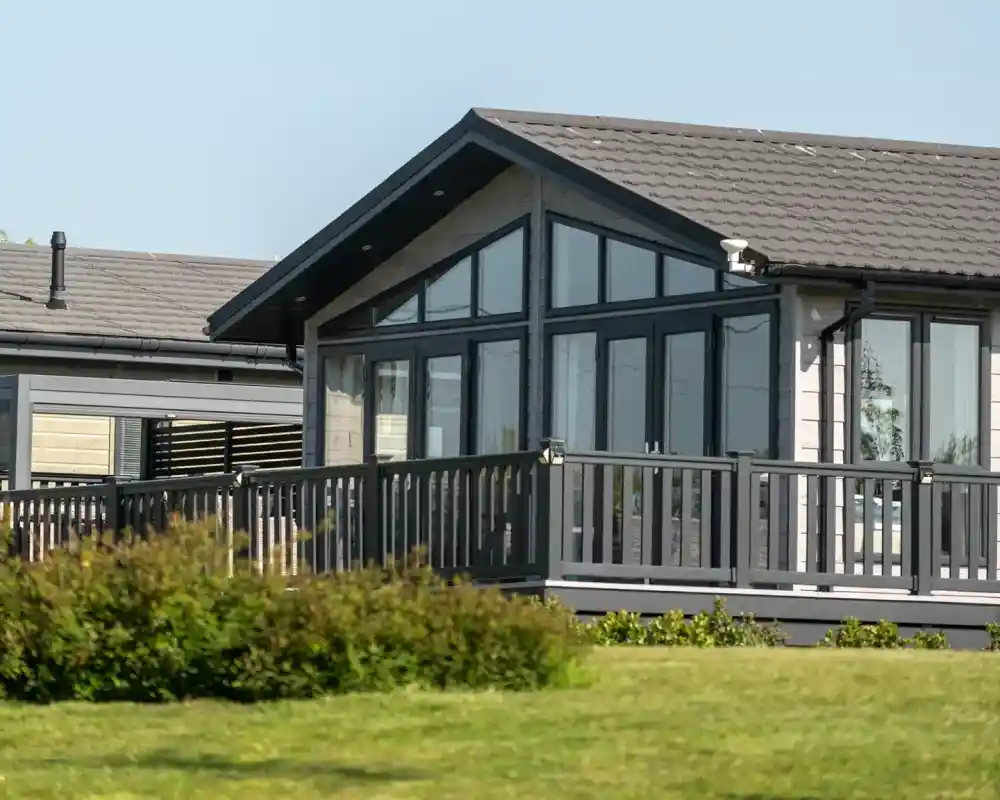 A modern house with large windows and a gray roof, featuring a spacious deck. Lush green grass surrounds the home.